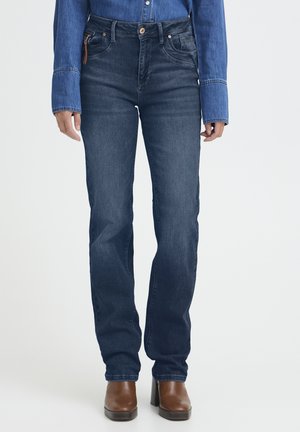 Mujer con jeans azul oscuro de pierna recta, botas marrones de tacón y una camisa vaquera de manga larga, posando frente a un fondo blanco.