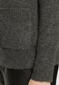 Grauer Wollpullover mit strukturiertem Strick, einer Taschenapplikation und einem leicht fransigen Saum. Der Stoff wirkt weich und warm.