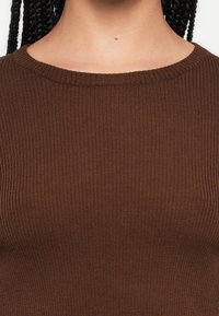 Hnedý rebrovaný pletený top s okrúhlym výstrihom. Materiál má jemnú textúru a zobrazuje horizontálne rebrovanie. Nie sú viditeľné žiadne ďalšie ozdoby.