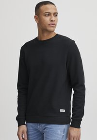 Schwarz strukturierter Sweatshirt mit rundem Hals, langen Ärmeln und gerippten Bündchen, versehen mit einem kleinen Etikett auf der unteren Seite. Getragen mit hellblauen Jeans.