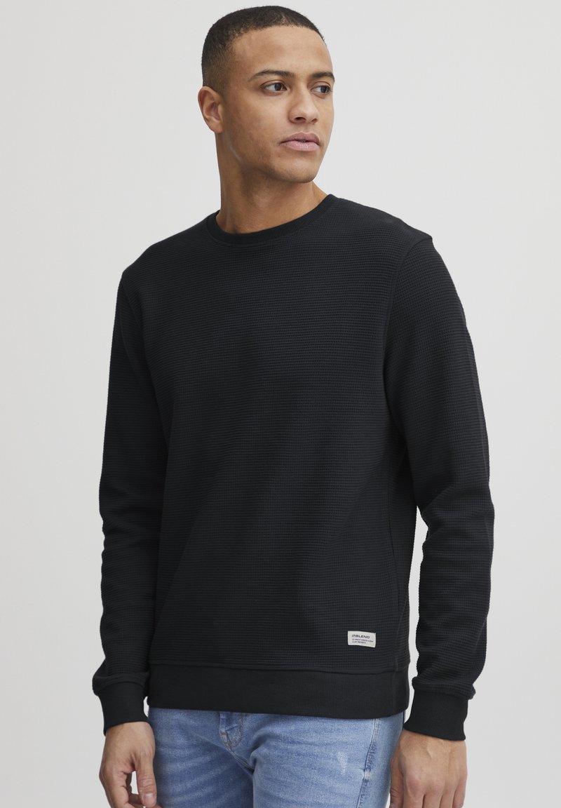 Schwarz strukturierter Sweatshirt mit rundem Hals, langen Ärmeln und gerippten Bündchen, versehen mit einem kleinen Etikett auf der unteren Seite. Getragen mit hellblauen Jeans.