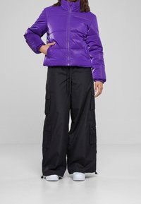 Fioletowa kurtka puchowa z wysokim kołnierzem i zamkiem, w zestawieniu z czarnymi szerokimi spodniami cargo z kieszeniami i sznurkiem. Białe sneakersy.