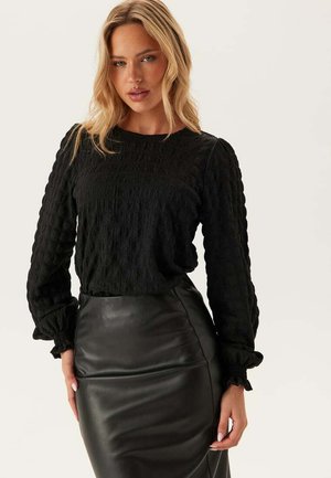 Blonde woman wearing textured black long-sleeve blouse and black leather pencil skirt, standing against a plain light background.