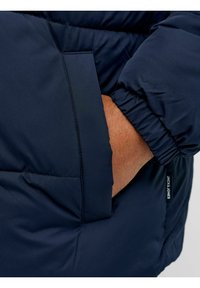 Marineblaue Daunenjacke mit glatter, gepolsterter Textur, elastischen Bündchen und einer Seitentasche mit markantem Label-Detail.