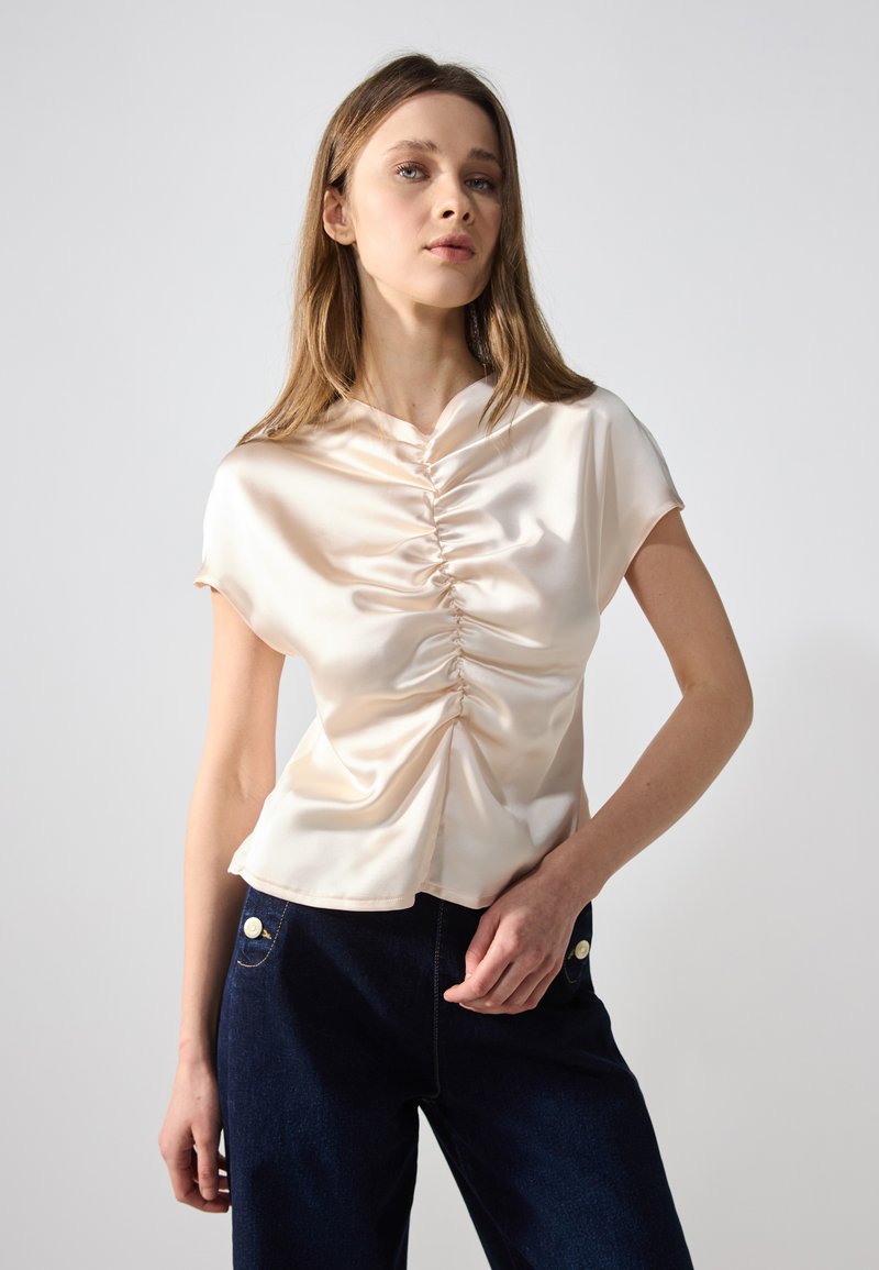 Top in satin a maniche corte in un morbido rosa pallido con dettaglio arricciato davanti, abbinato a pantaloni in denim scuro a vita alta con bottoni di perla.