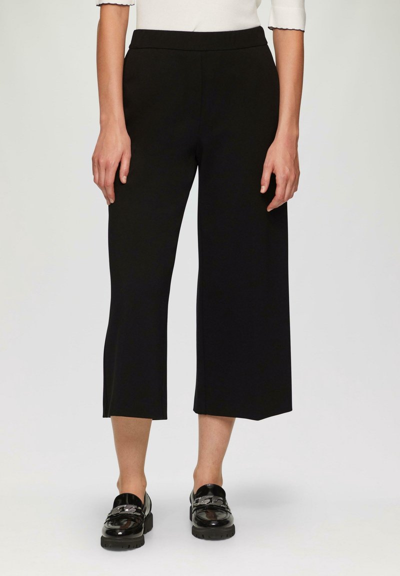 Pantalon noir à jambes larges et courtes avec une texture lisse et une taille élastique, assorti à des mocassins noirs brillants ornés d'accents chaîne.