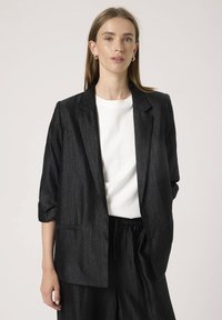 Čierny textúrovaný blazer s uvoľneným strihom, zatočenými rukávmi, vykrojeným golierom a jednou prednou vreckom, nosený cez biele tričko.