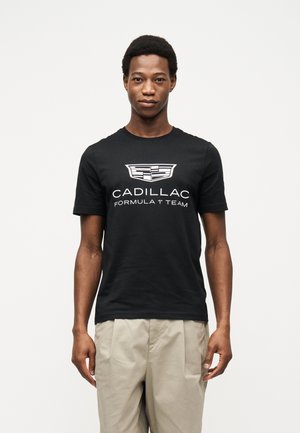 Joven de pie mirando al frente, vistiendo una camiseta negra del "Equipo de Fórmula 1 Cadillac" y pantalones beige, con un fondo neutro.