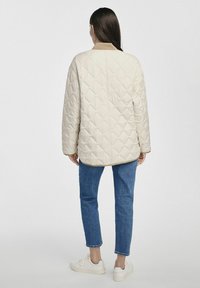 Jasno beżowa pikowana kurtka z wysokim kołnierzem, wzorem w romby i luźnym krojem. Połączona z niebieskimi dżinsami i białymi trampkami.