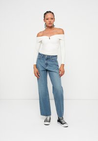 White off-the-shoulder top with a button detail, paired with light blue wide-leg jeans and black high-top sneakers. Plain background.