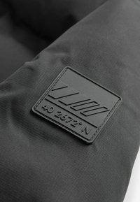 Tissu noir avec une surface texturée et un patch en cuir noir carré embossé avec "40 2672° N" et des lignes géométriques.
