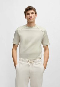 Camicia a maniche corte beige chiaro con finitura testurizzata, colletto rotondo e pannelli delle spalle lisci a contrasto, abbinata a pantaloni di colore chiaro.