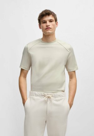 Camicia a maniche corte beige chiaro con finitura testurizzata, colletto rotondo e pannelli delle spalle lisci a contrasto, abbinata a pantaloni di colore chiaro.