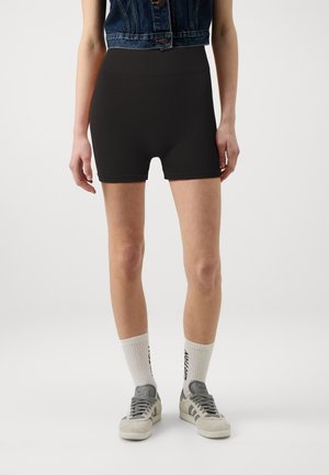 Personne portant un short de vélo noir taille haute, des chaussettes blanches avec texte noir, des baskets grises et blanches, et un gilet en jean sans manches.