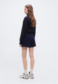 Γυναίκα με μακριά καστανά μαλλιά, φορώντας μαύρο μπλουζάκι μακρυμάνικο, σκούρο μπλε σορτς, άσπρες κάλτσες και άσπρα αθλητικά παπούτσια, κοιτάζει προς τα αριστερά σε απλό φόντο.