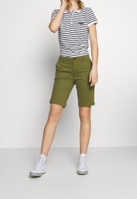 Vihreät puuvillahousut, joissa käärityt lahkeet, yhdistetty tummansinivalkoraidalliseen t-paitaan. Malli pukeutunut valkoisiin tennareihin, kädet lähellä kasvoja.