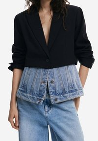 Schwarzer, kurz geschnittener Blazer mit einem unteren Abschnitt aus Denim; verfügt über eine Knopfverschluss und ein geschichtetes Design. Getragen über hellblauen Denim-Hosen.