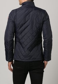 Marineblau gesteppte Jacke mit Stehkragen, diagonalen Nähten, Seitentaschen und schlanker Passform, kombiniert mit schwarzen Hosen vor einem neutralen Hintergrund.