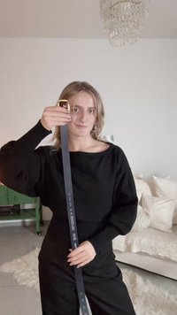 Mustavöinen vyö, jossa on kullanvärinen solki, näyttää pituuden ja bränditekstin. Malli käyttää mustaa yläosaa, joka esittelee rentoa istuvuutta ja tekstuuria. Valkoiset tyynyt taustalla.