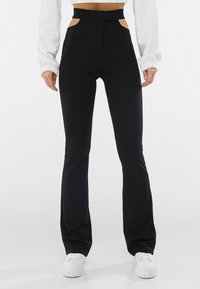 Höga svarta byxor med skurna sidor, en figurnära design, utsvängda ben och en slät textur, parat med vita sneakers.