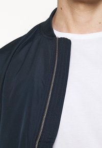 Námořnicky modrá bomber bunda s hladkým povrchem, žebrovaným límcem a stříbrným zipem. Nasažená přes bílé tričko, zdůrazňující vrstvení.