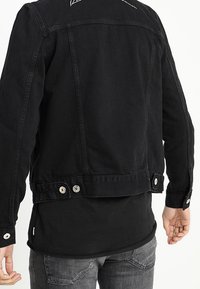 Černá džínová bunda s vysokým límcem, bílým prošíváním na ramenou a knoflíkovými ozdobami na lemu. Hladká, pevná textura.