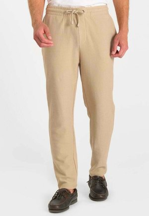 Mann trägt beige strukturierte Jogginghosen mit Kordelzug und dunkelbraune Mokassins, steht vor weißem Hintergrund.