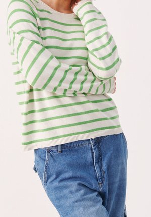 Person iført en hvid sweater med lysegrønne vandrette striber og blå jeans, stående med armene krydset og hånden tæt på ansigtet.