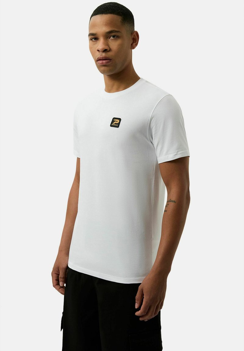 Weißes kurzärmliges T-Shirt aus glattem Stoff mit einem kleinen, schwarzen und goldenen Logo-Patch auf der Brust. Getragen mit schwarzen Hosen.