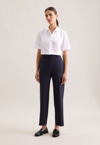 Weißes Hemd mit kurzen Ärmeln und Knöpfen sowie marineblaue, maßgeschneiderte Hosen mit leichten Falten. Modell trägt schwarze Loafer. Neutraler Hintergrund.