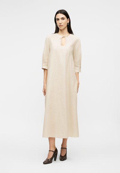 Γυναίκα με ίσια σκούρα μαλλιά φοράει μπεζ μακρύ φόρεμα με μανίκια 3/4, άνοιγμα στο μπροστινό μέρος και κορδόνι, σε συνδυασμό με σκουρόκαφέ ψηλοτάκουνα παπούτσια.