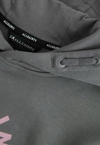 Sivá mikina s kapucňou so šnúrkami a čiernymi štítkami značky AllSaints vo vnútri goliera, zobrazujúca časť ružového tlačeného textu na látke.