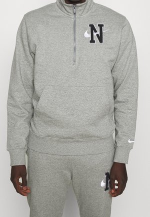 Person iført en grå Nike træningsdragt med sort bogstav "N" og hvide swoosh-logoer på brystet, benet og venstre håndled.