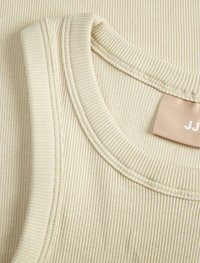 Lyse beige ribbet tanktopstof med en rund halsudskæring og synlige sømdetaljer; et lille mærke er delvist synligt.
