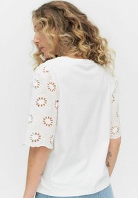 Vit t-shirt med korta ärmar som har blommig broderad utskärning och vågiga kanter. Slät textur med en avslappnad design.