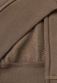 Tessuto di felpa marrone con texture a costine, fodera in pile morbido e dettaglio di cucitura sul bordo. Il materiale sembra caldo e accogliente.