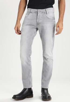 Hombre que lleva jeans ajustados de color gris claro y botines Chelsea de cuero negro, de pie contra un fondo blanco.