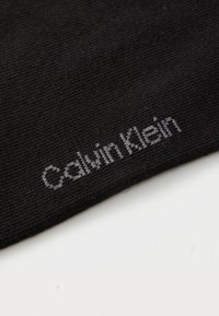 Schwarzer Stoff mit weißem, nahe dem Rand gesticktem "Calvin Klein"-Logo auf einer glatten, hellen Oberfläche.