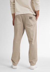 Beige katoenen broek met een relaxte pasvorm, twee achterzakken en een logo patch. Heeft elastische boorden en wordt gedragen met witte sneakers.