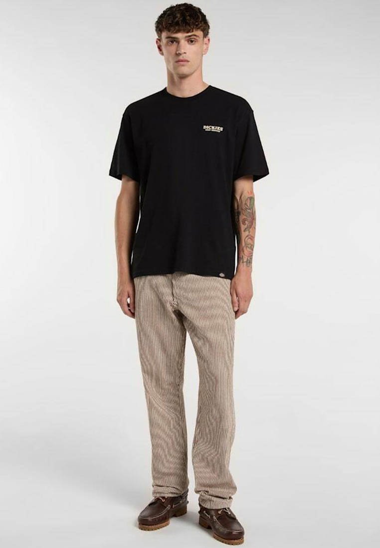 Ung mand med kort krøllet hår iført en sort Dickies t-shirt, beige stribede bukser, brune sko og tatovering på venstre underarm.