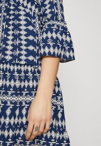 Blåmønstret kjole med et hvitt geometrisk design, flagrende ermer og en tilpasset midje. Hånd med synlig tatovering hviler på stoffet.