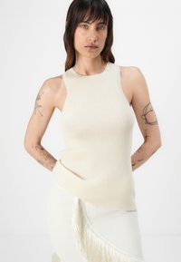 Top tank côtelé beige avec un col haut et sans manches. Texture douce, associé à une jupe blanche avec des détails à franges.