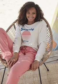 Crème sweatshirt met blauwe "Sunrise"-tekst en grafiek, gecombineerd met zachte roze joggers met trekkoorddetail; zittend in een geweven stoel.