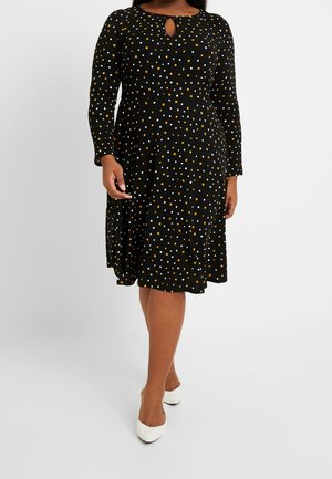 Vrouw draagt een zwarte jurk tot op de knie met witte en oranje polkadots, lange mouwen, een keyhole-halslijn en witte puntige schoenen.