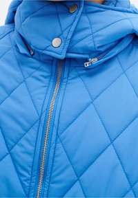 Chaqueta acolchada azul con un alto cuello, cremallera metálica y botones de presión. La tela es suave con un ligero brillo. Detalle de patrón en diamante.