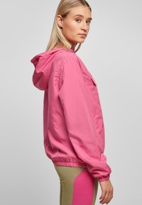 Lichtgewicht roze jas met een capuchon, elastische manchetten en een zak. Vlotte textuur en een relaxte pasvorm, gedragen over beige en roze leggings.