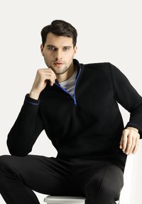 Sudadera negra con cremallera, hecha de un tejido suave, que presenta una cremallera y detalles en azul contrastante, con un ajuste relajado y mangas largas.