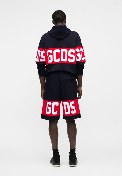 Άνδρας που φοράει μαύρη και κόκκινη μπλούζα με κουκούλα και ταιριαστά σορτς με μεγάλα λευκά γράμματα GCDS στην πλάτη και στο κάτω μέρος των σορτς.