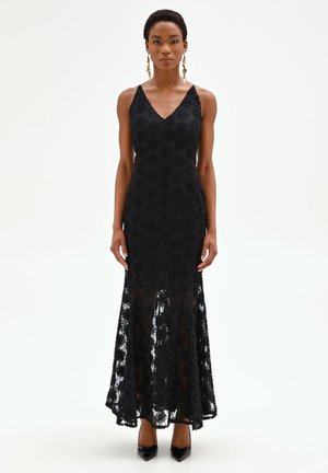 Femme portant une robe en dentelle noire sans manches avec un col en V et un ourlet transparent, associée à des talons pointus noirs et des boucles d'oreilles dorées.