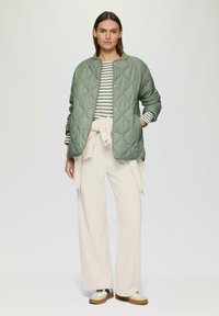Veste verte matelassée, chemise à rayures à manches longues, pantalon blanc ajusté, léger pull noué à la taille, chaussures décontractées avec des accents marron.